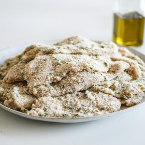Plate of breaded chicken schnitzel, ready for cooking, with olive oil bottle in background.